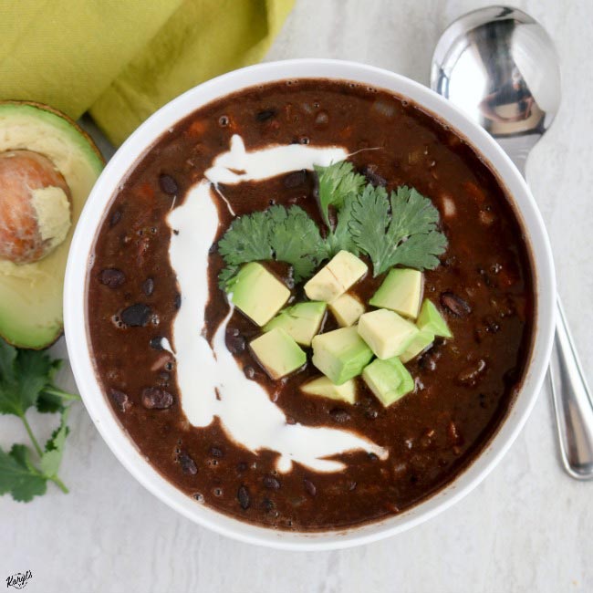 overhead shot of a white bowl with black bean soup with half an avocado and a metal spoon on the side