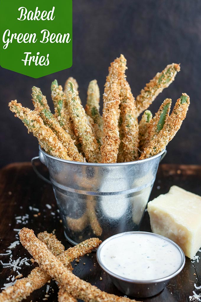 close up shot of Baked Green Bean Fries in a metal bucket with a side of ranch in front and a block of Parmesan cheese on the side