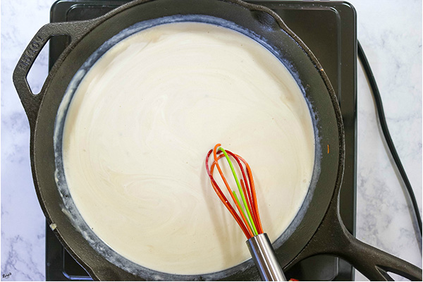 overhead process shot: milk, broth and heavy cream mixed into the roux in a black skillet, with a whisk on the side