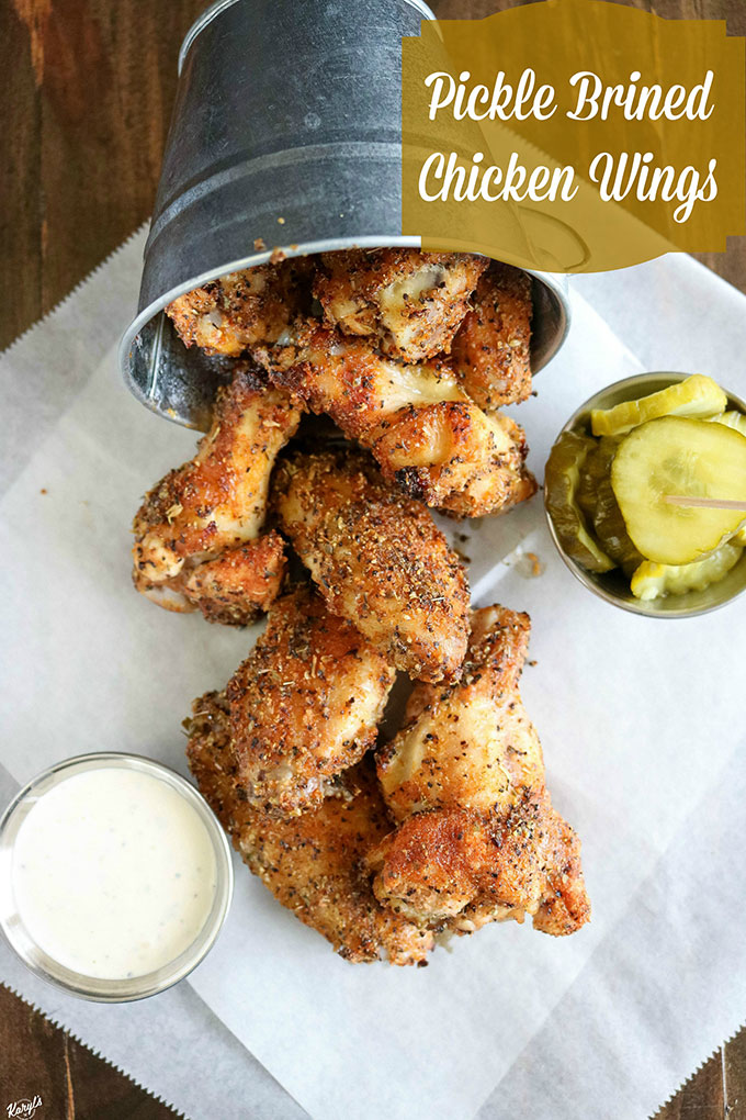 overhead shot of finished chicken wings falling out of a metal bucket onto white paper, with dip and pickles on the side