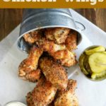 overhead shot of finished chicken wings falling out of a metal bucket onto white paper, with dip and pickles on the side