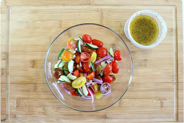 overhead process shot: tomatoes, onion & cucumbers in a glass bowl with a container of dressing on the side